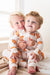 Two young children sitting together indoors, wearing matching long-sleeve footless pajamas with an orange teddy bear print on a cream background.