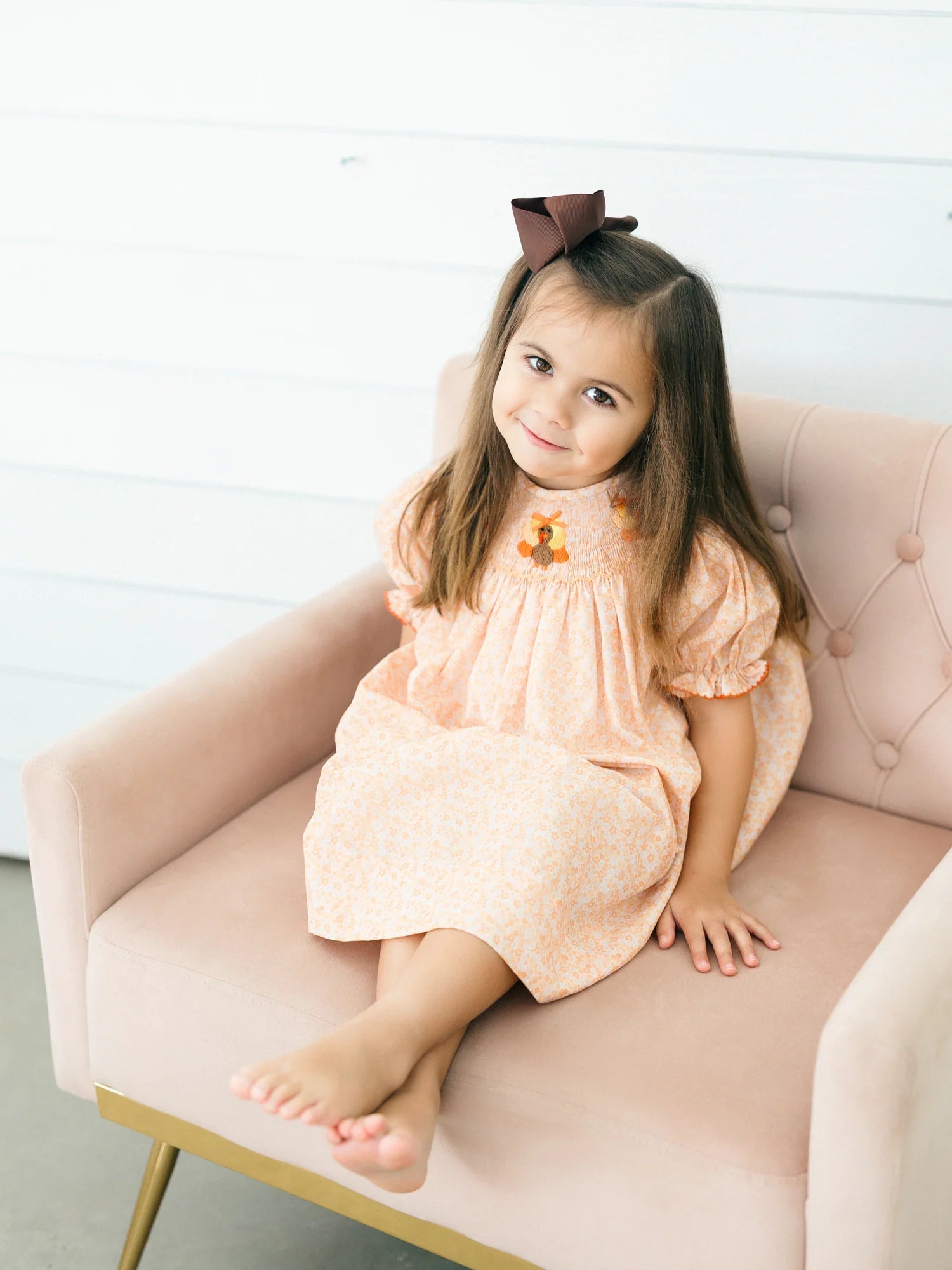 Young girl wearing a peach floral dress with puff sleeves and a turkey detail while sitting barefoot in a blush pink chair with a brown bow in her hair for Poppy Kids