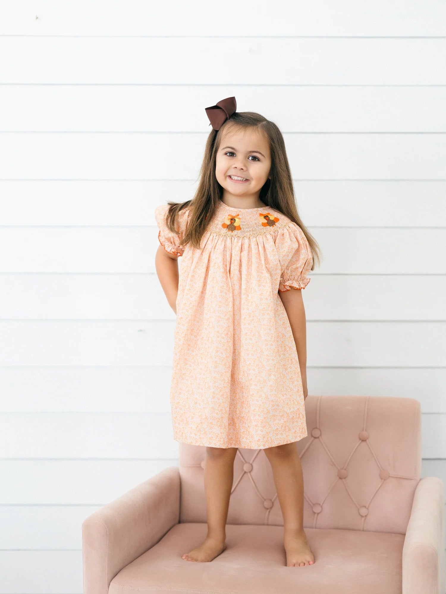Young girl wearing a peach floral dress with turkey embroidery on the smocked yoke and puff sleeves while standing on a blush pink chair for Poppy Kids