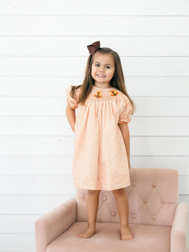 Young girl wearing a peach floral dress with turkey embroidery on the smocked yoke and puff sleeves while standing on a blush pink chair for Poppy Kids
