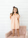 Young girl wearing a peach floral dress with turkey embroidery on the smocked yoke and puff sleeves while standing on a blush pink chair for Poppy Kids