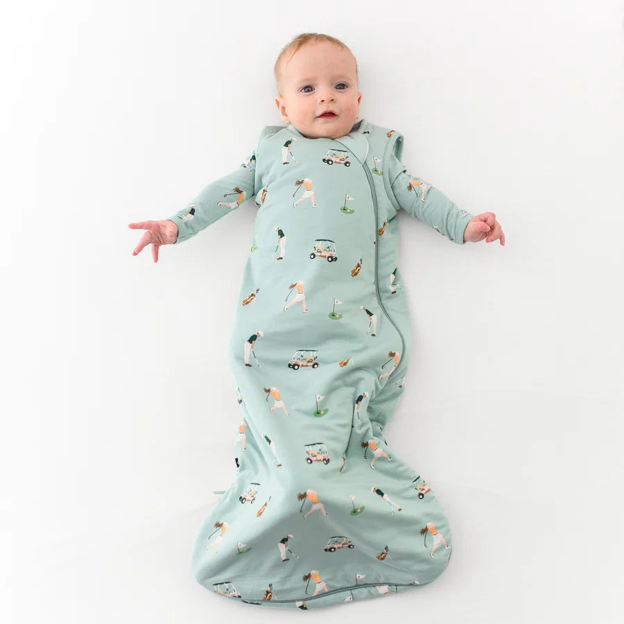 A baby lying on their back wearing a mint green long-sleeve sleep sack with a golf print, set against a plain white background.