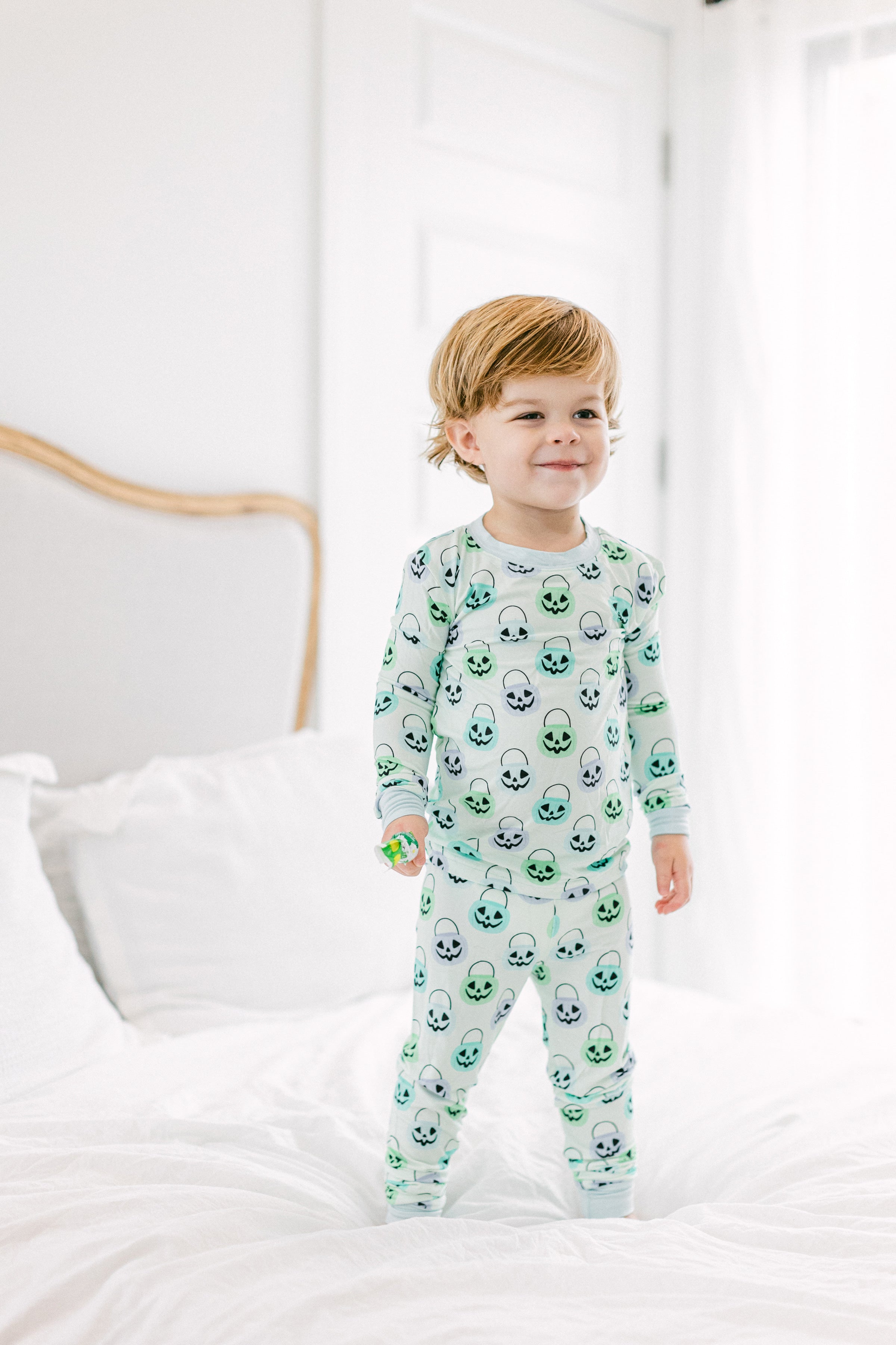 A toddler stands on a white bed wearing a light green and white long-sleeve pajama set with an all-over Halloween pumpkin print. The background shows a softly lit bedroom with white bedding and a neutral headboard.