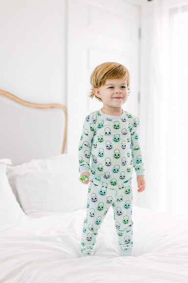 A toddler stands on a white bed wearing a light green and white long-sleeve pajama set with an all-over Halloween pumpkin print. The background shows a softly lit bedroom with white bedding and a neutral headboard.