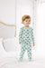 A toddler stands on a white bed wearing a light green and white long-sleeve pajama set with an all-over Halloween pumpkin print. The background shows a softly lit bedroom with white bedding and a neutral headboard.