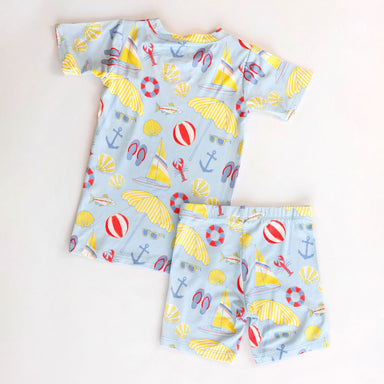 Children's pajamas with a blue pattern featuring yellow umbrellas, red life buoys, and white clouds on a light beige background.