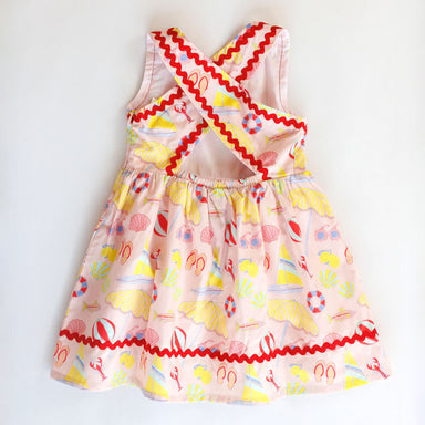 Colorful children's dress with a pattern of flip-flops, seashells, and other beach-themed elements on a white background.
