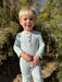 A young toddler with blonde hair stands outdoors in front of green bushes, wearing a light heather gray long-sleeve henley jumpsuit with three brown buttons.