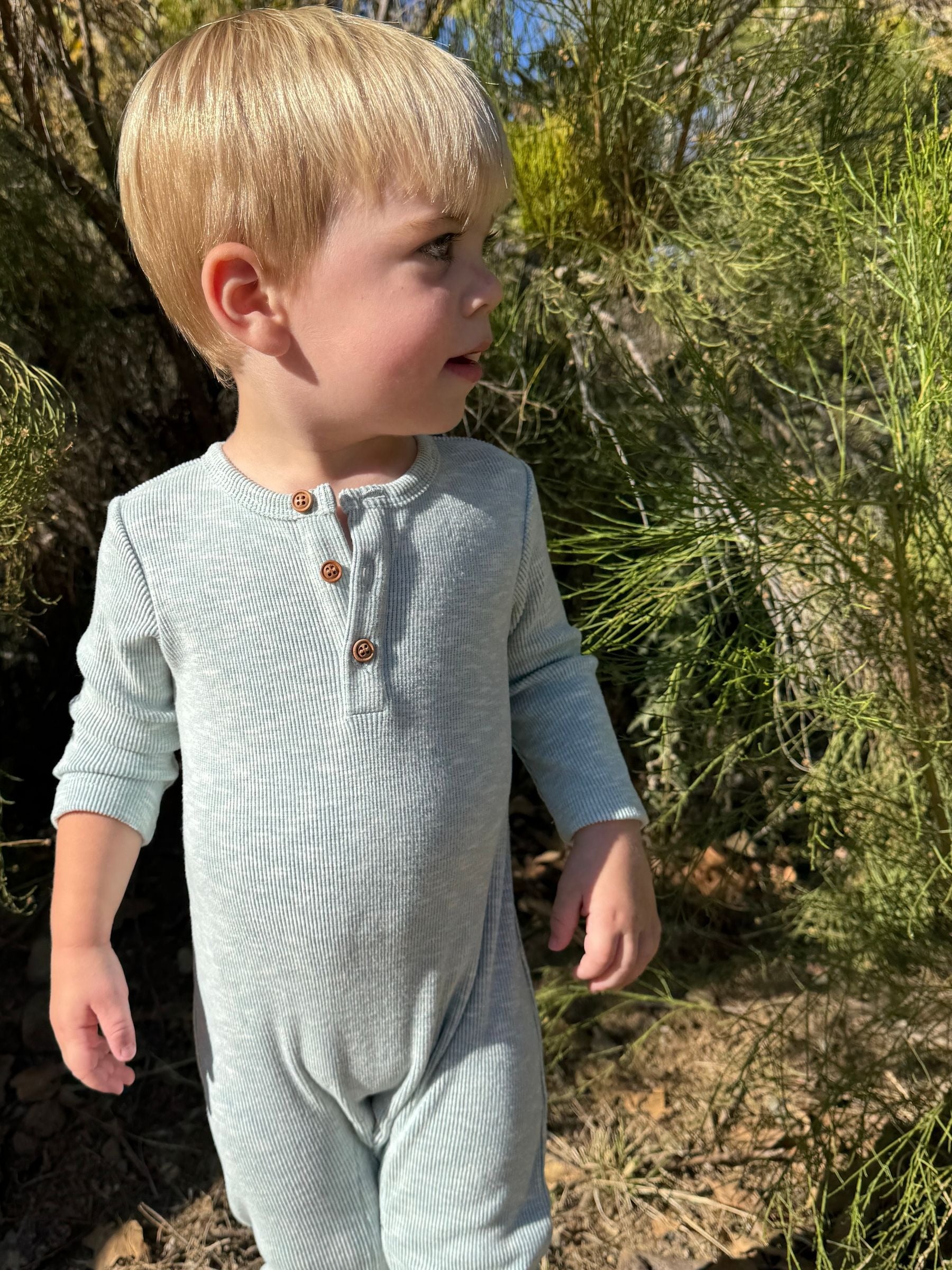A young blonde toddler wearing a light gray ribbed long-sleeve henley jumpsuit, standing outdoors in front of green shrubs.