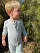 A young blonde toddler wearing a light gray ribbed long-sleeve henley jumpsuit, standing outdoors in front of green shrubs.