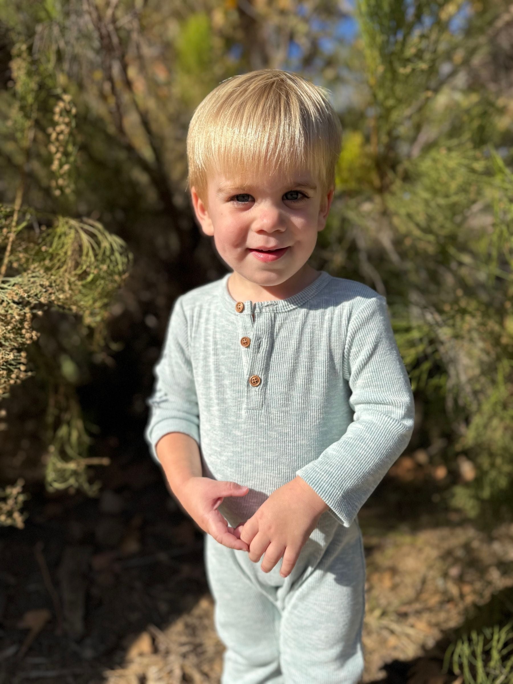 A young toddler with blonde hair wearing a light sage green ribbed long-sleeve henley jumpsuit, standing outdoors in front of green bushes.