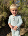 A young toddler with blonde hair wearing a light sage green ribbed long-sleeve henley jumpsuit, standing outdoors in front of green bushes.