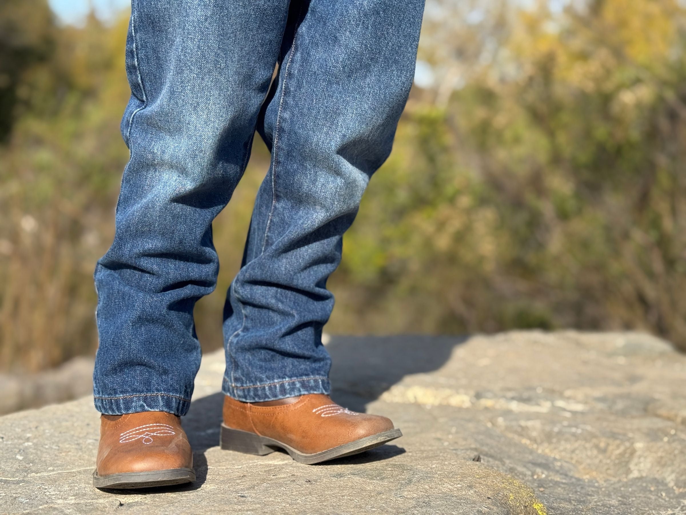 A person wearing blue jeans and brown leather western-style boots standing outdoors on a rock with a blurred natural background.