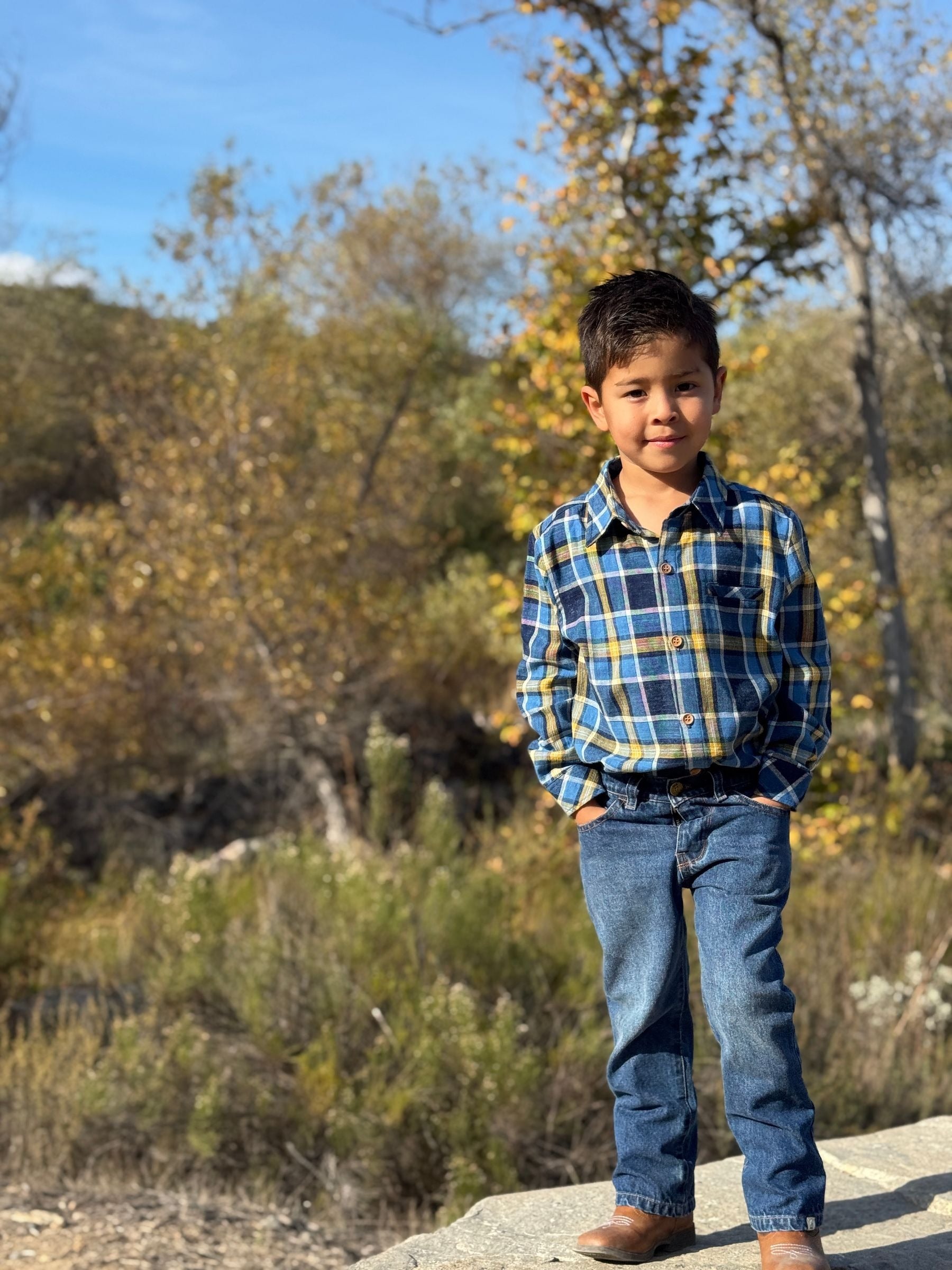 Child wearing a plaid shirt and jeans standing outdoors with trees and shrubs in the background