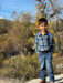 Child wearing a plaid shirt and jeans standing outdoors with trees and shrubs in the background