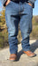 A person wearing medium-wash blue denim jeans and brown leather cowboy boots, standing outdoors on a stone surface with trees in the background.