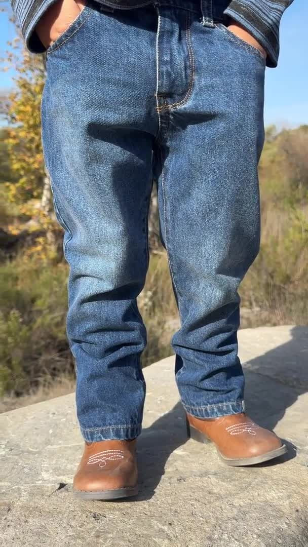 A person wearing medium-wash blue denim jeans and brown leather cowboy boots, standing outdoors on a stone surface with trees in the background.