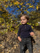 A toddler standing outdoors in a wooded area, wearing a dark navy long-sleeve henley shirt with brown buttons and dark gray jeans.