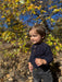 A toddler boy wearing a navy blue long-sleeve henley shirt and dark gray jeans, standing outdoors in a wooded area with autumn leaves and blue sky in the background.