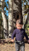 A toddler wearing a dark purple long-sleeve henley shirt and dark gray jeans walking outdoors in front of a large tree trunk.