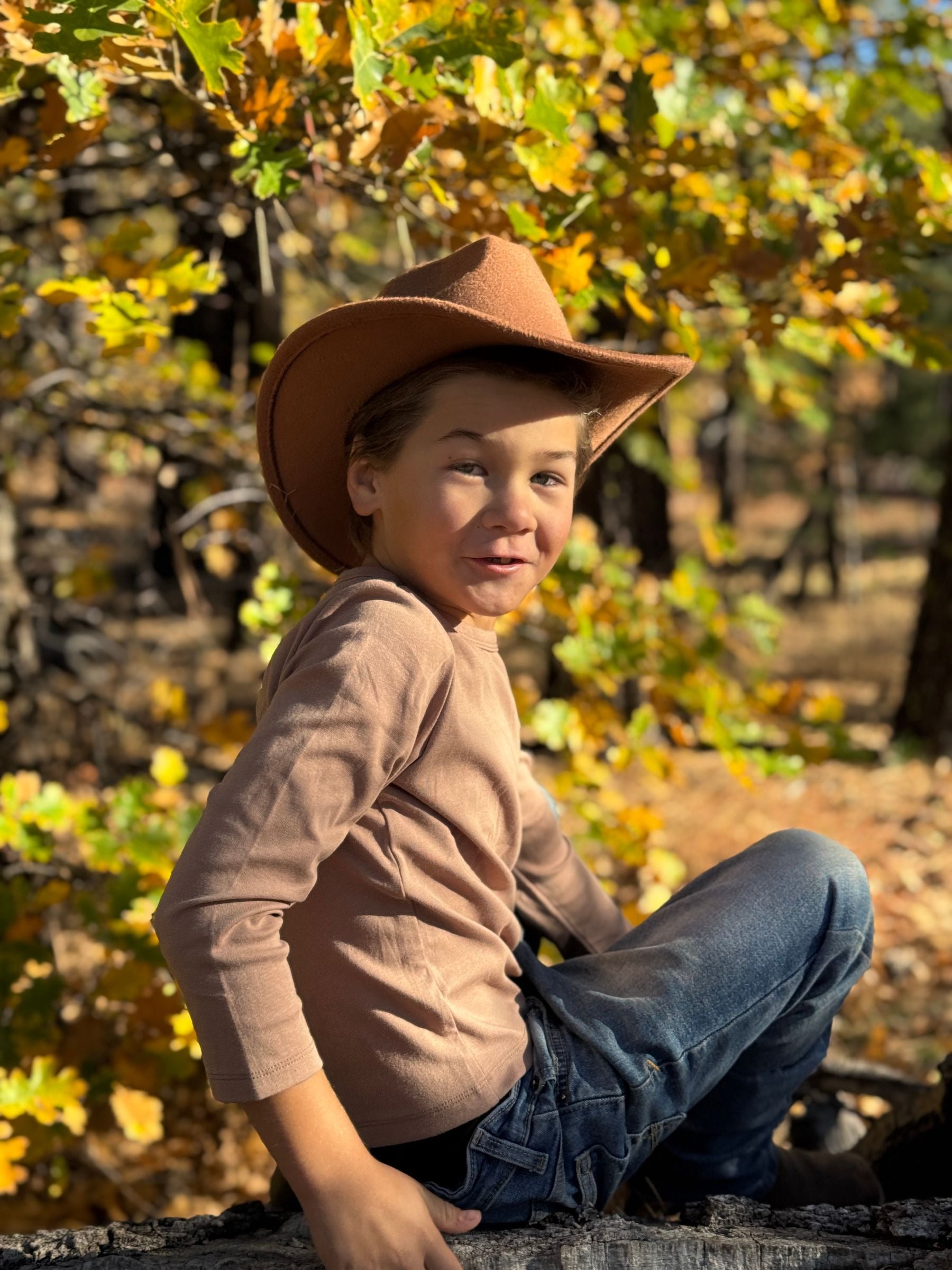 Child wearing a cowboy hat sitting among trees with autumn leaves