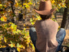 A person seen from behind wearing a tan cowboy hat, a light beige graphic tank top with 'I SHOULDA BEEN TX A COWBOY' text, and blue jeans, sitting outdoors in a wooded area with autumn foliage.