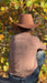 A person seen from behind wearing a brown cowboy hat, a dusty pink graphic t-shirt with 'COWBOY' printed on the back, and blue jeans, sitting outdoors among autumn trees with yellow and orange leaves.