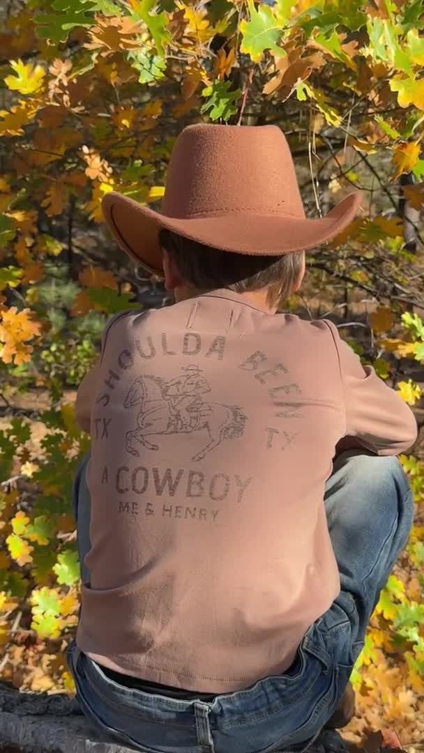 A person seen from behind wearing a brown cowboy hat, a dusty pink graphic t-shirt with 'COWBOY' printed on the back, and blue jeans, sitting outdoors among autumn trees with yellow and orange leaves.
