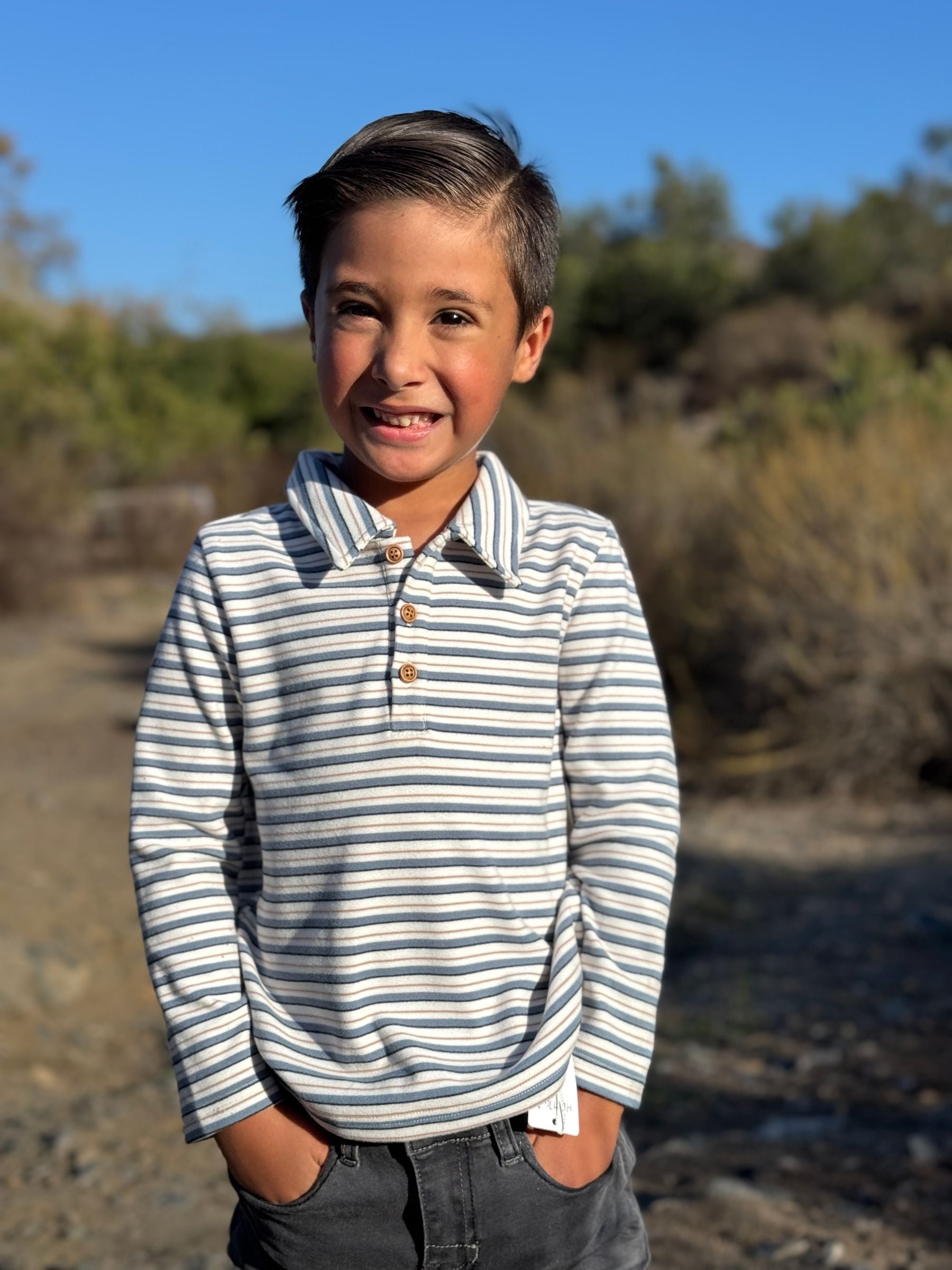 A young boy smiling outdoors, wearing a long-sleeve polo shirt with thin navy and off-white horizontal stripes, paired with dark gray jeans.