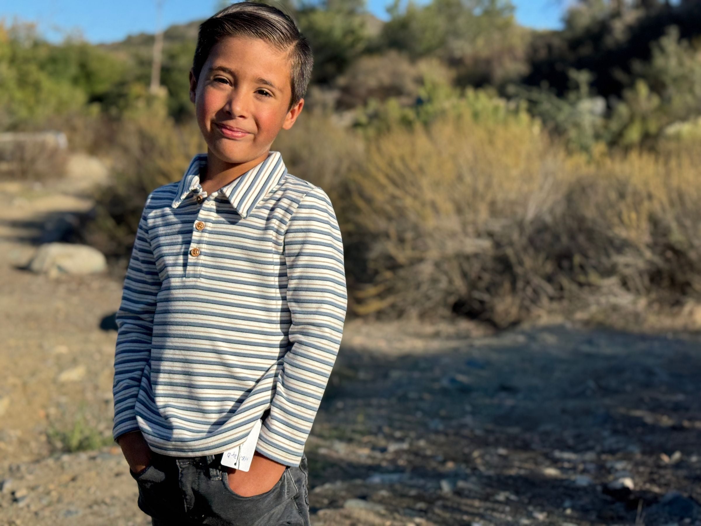A young boy smiling outdoors, wearing a long-sleeve polo shirt with thin blue and white horizontal stripes and dark gray pants, standing in a sunlit natural setting with dry brush and trees in the background.