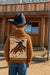 Child wearing a brown sweater with a cowboy design, standing in front of a wooden building.