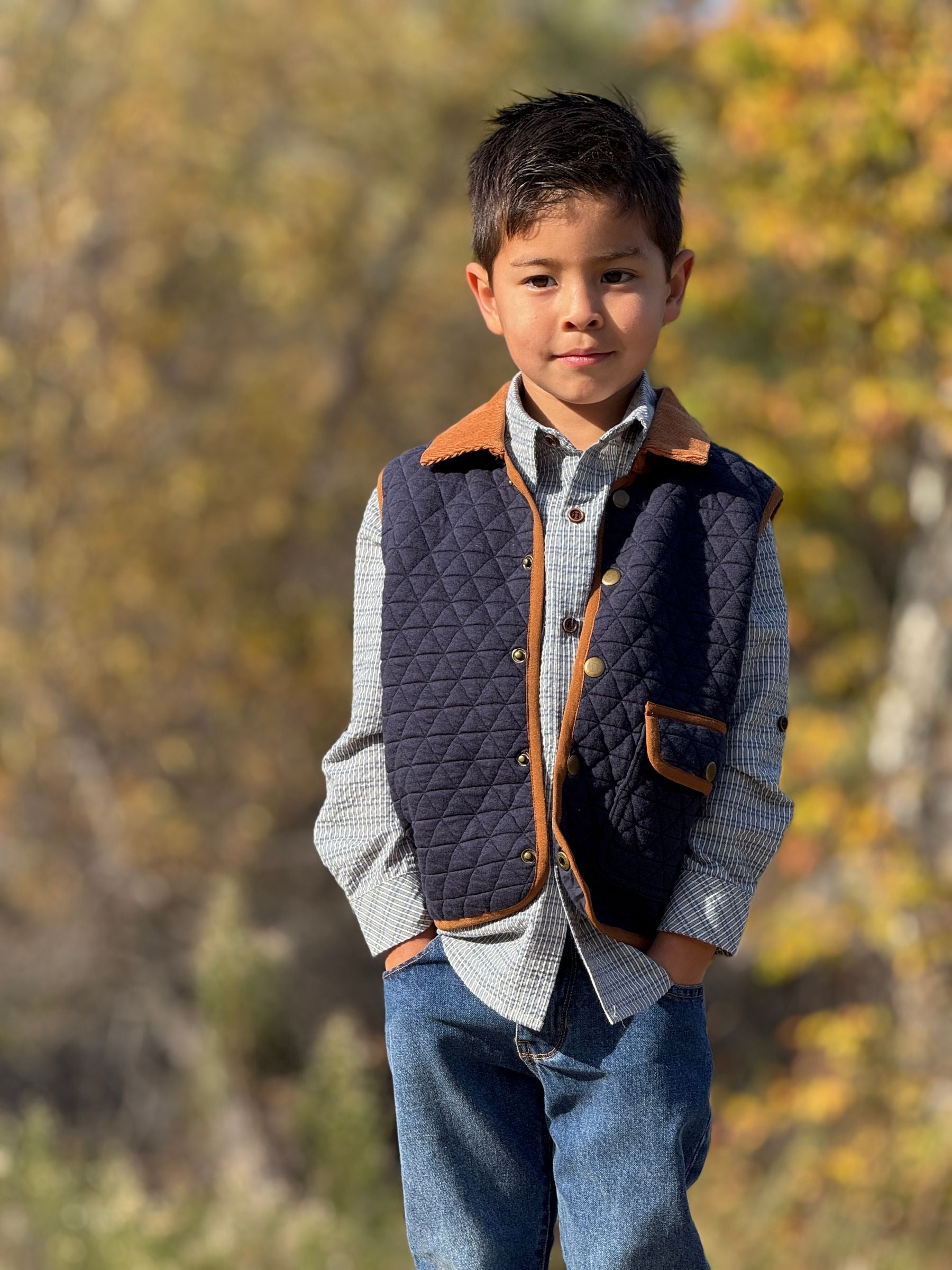 A young boy wearing a quilted navy blue vest with brown corduroy trim over a light blue and white checkered shirt and blue jeans, standing outdoors against a blurred autumn background.