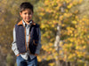 A young boy wearing a quilted navy blue vest with brown trim over a light blue and white checkered shirt and blue jeans, standing outdoors against a blurred background of yellow and green autumn trees.