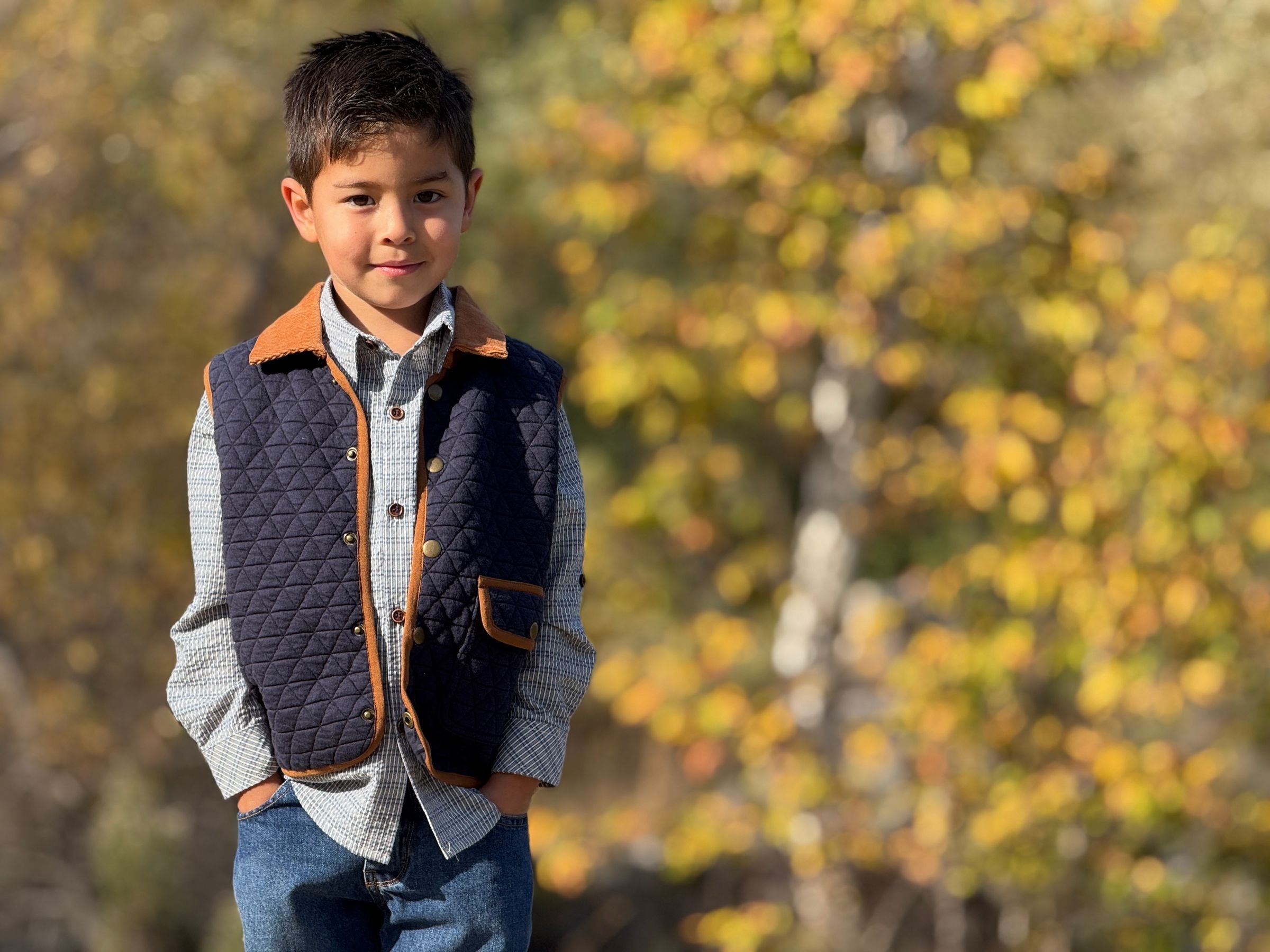 A young boy wearing a quilted navy blue vest with brown trim over a light blue and white checkered shirt and blue jeans, standing outdoors against a blurred background of yellow and green autumn trees.