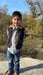 A young boy stands outdoors on a stone surface wearing a quilted navy vest with brown trim over a light patterned button-up shirt and blue jeans, with trees and blue sky in the background.