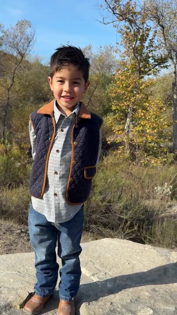 A young boy stands outdoors on a stone surface wearing a quilted navy vest with brown trim over a light patterned button-up shirt and blue jeans, with trees and blue sky in the background.