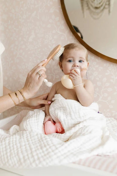 Baby being bathed with a brush and soap, wrapped in a towel on a soft surface.