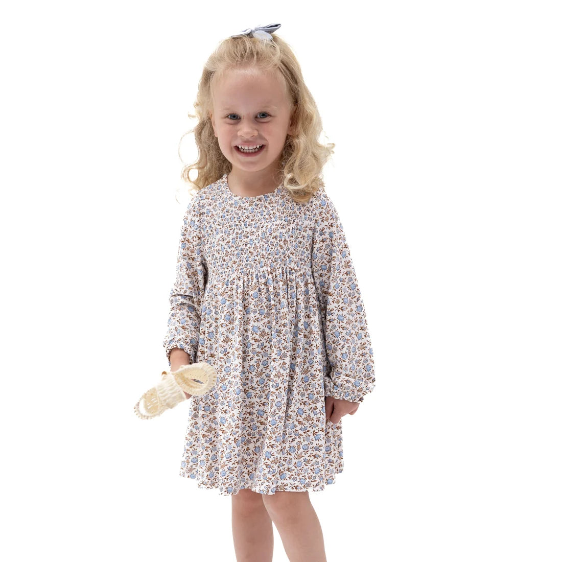 A young girl with blonde curly hair wearing a long-sleeve babydoll dress with a small multicolor floral pattern, holding a small cream-colored bootie against a plain white background.