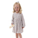 A young girl with blonde curly hair wearing a long-sleeve babydoll dress with a small multicolor floral pattern, holding a small cream-colored bootie against a plain white background.