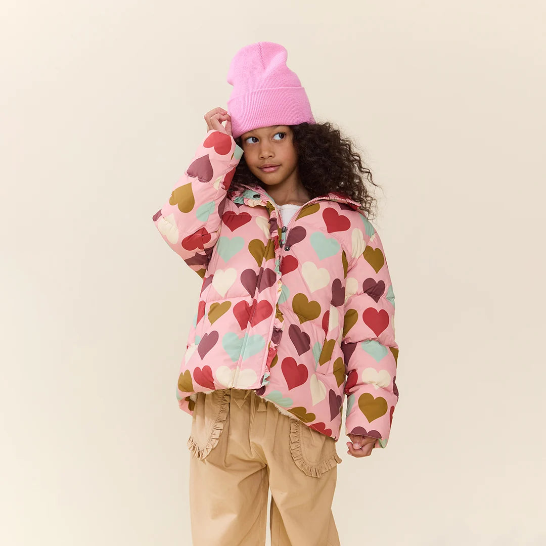 A young girl with curly dark hair wears a pink knit beanie and a pink puffer jacket covered in colorful heart prints, photographed against a plain light beige background.