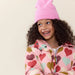 A smiling young girl with curly dark hair wears a pink knit beanie and a pink puffer jacket covered in colorful heart prints, photographed against a plain light beige background.