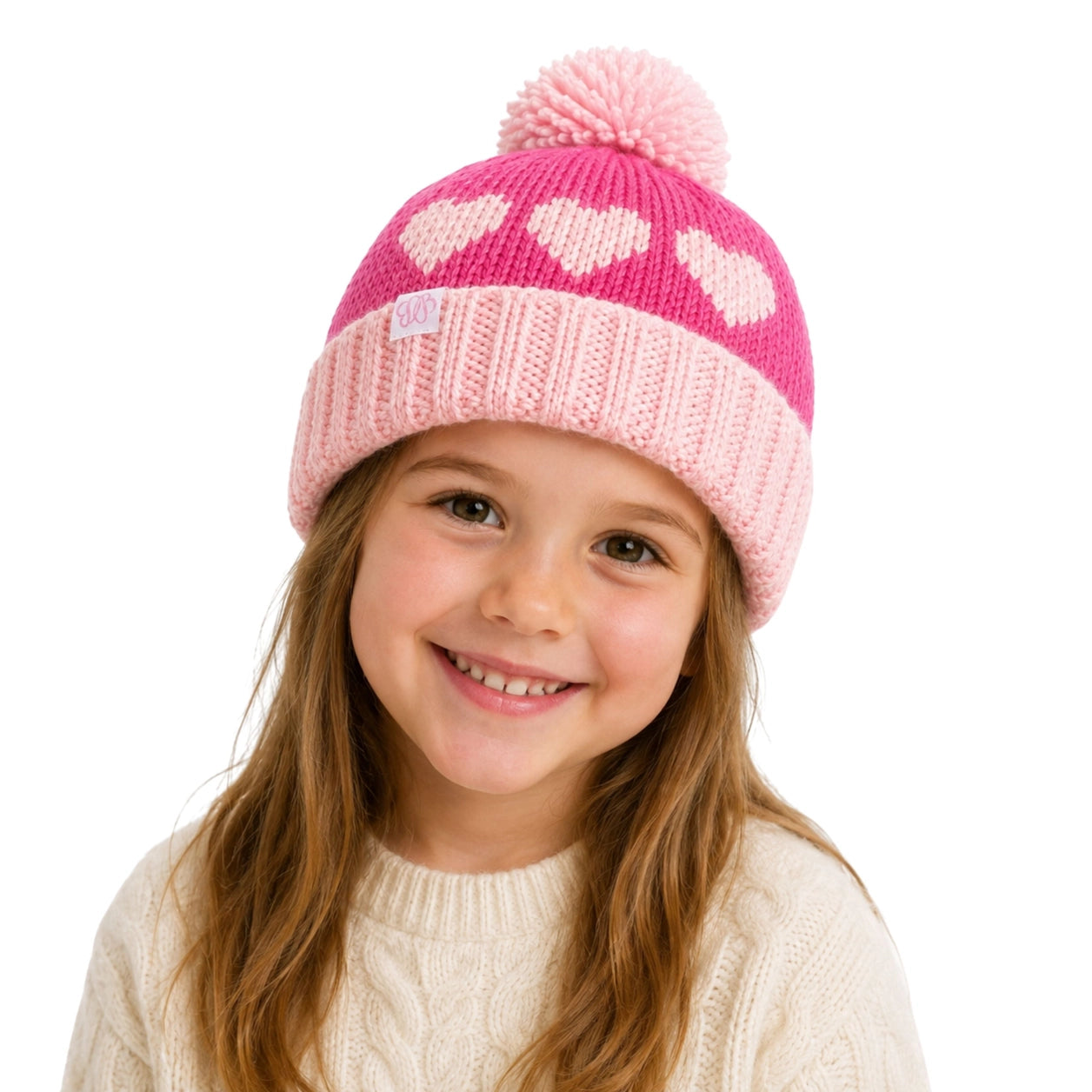 A young girl with long light brown hair smiling while wearing a pink and light pink knit beanie with heart patterns and a pom-pom, paired with a cream cable-knit sweater against a solid white background.