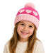 A young girl with long light brown hair smiling while wearing a pink and light pink knit beanie with heart patterns and a pom-pom, paired with a cream cable-knit sweater against a solid white background.