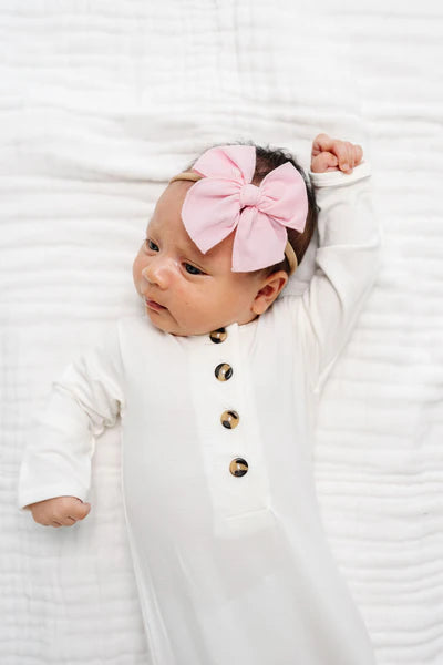 Newborn baby wearing a white outfit with gold buttons and a pink bow headband on a white background