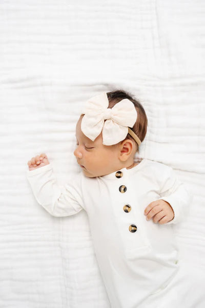Newborn baby wearing a white outfit with gold buttons and a large bow headband on a white background