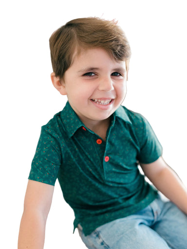 A young boy smiling and looking to the side, wearing a dark green short-sleeve polo shirt with tiny holly pattern and red buttons, paired with light blue jeans.