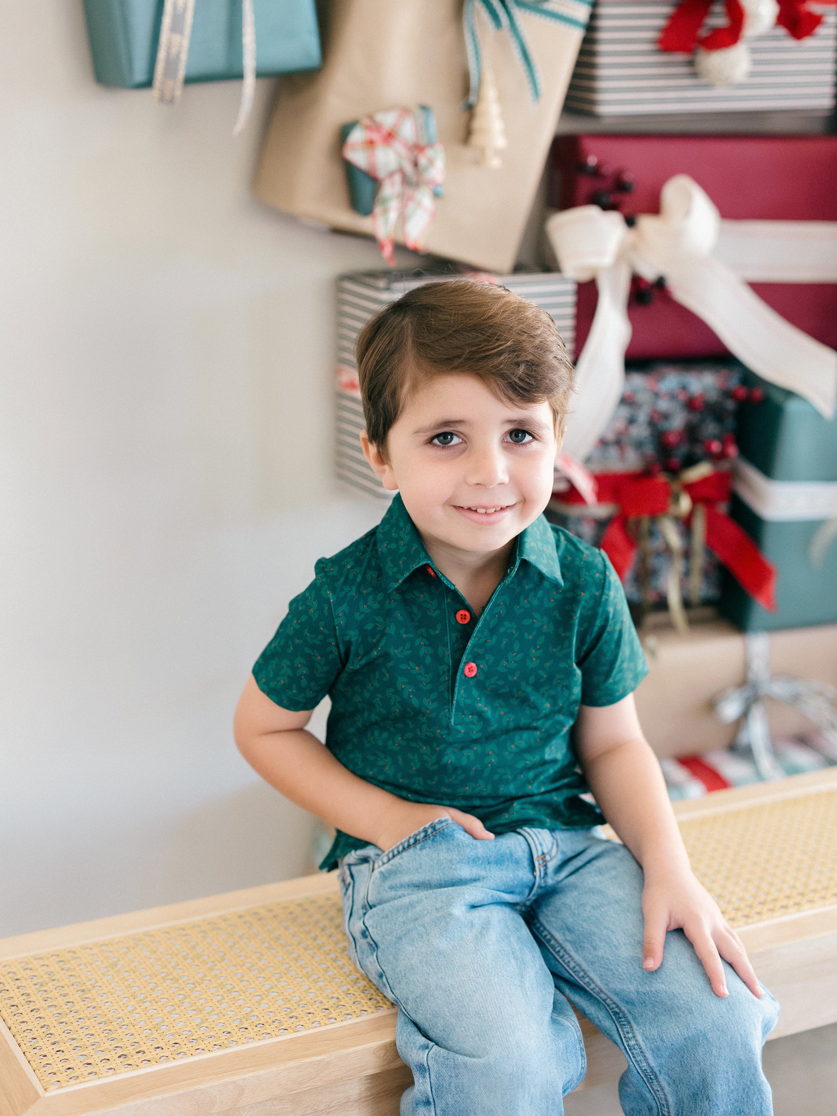 A young boy with brown hair sits on a woven bench, wearing a dark green short-sleeve button-up shirt with a subtle holly pattern and light blue jeans. The background features a soft-focus holiday setting with wrapped gifts and hanging paper bags.