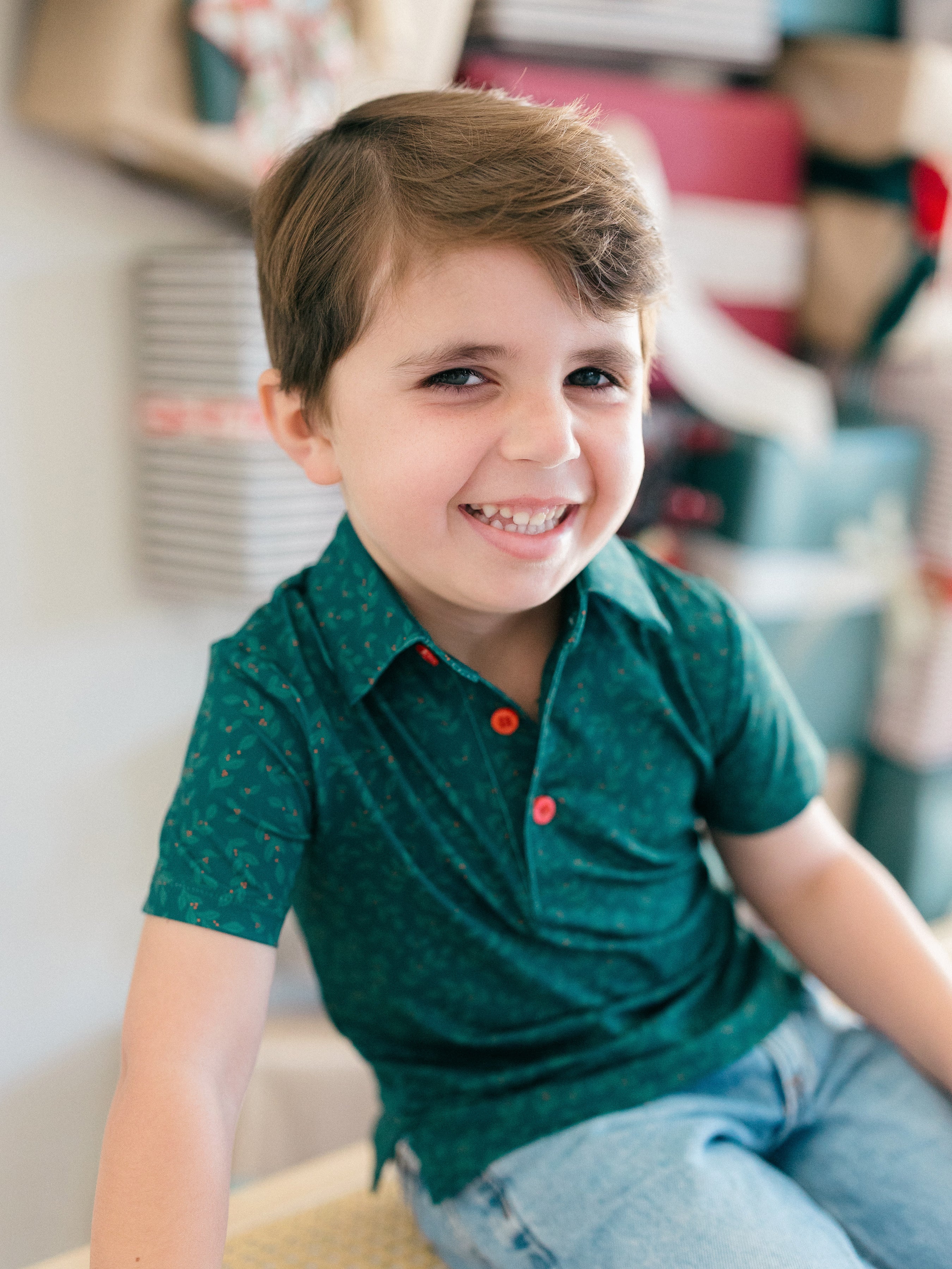 A young boy smiling and looking to the side, wearing a dark green short-sleeve polo shirt with tiny holly pattern and red buttons, paired with light blue jeans.