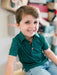A young boy smiling and looking to the side, wearing a dark green short-sleeve polo shirt with tiny holly pattern and red buttons, paired with light blue jeans.
