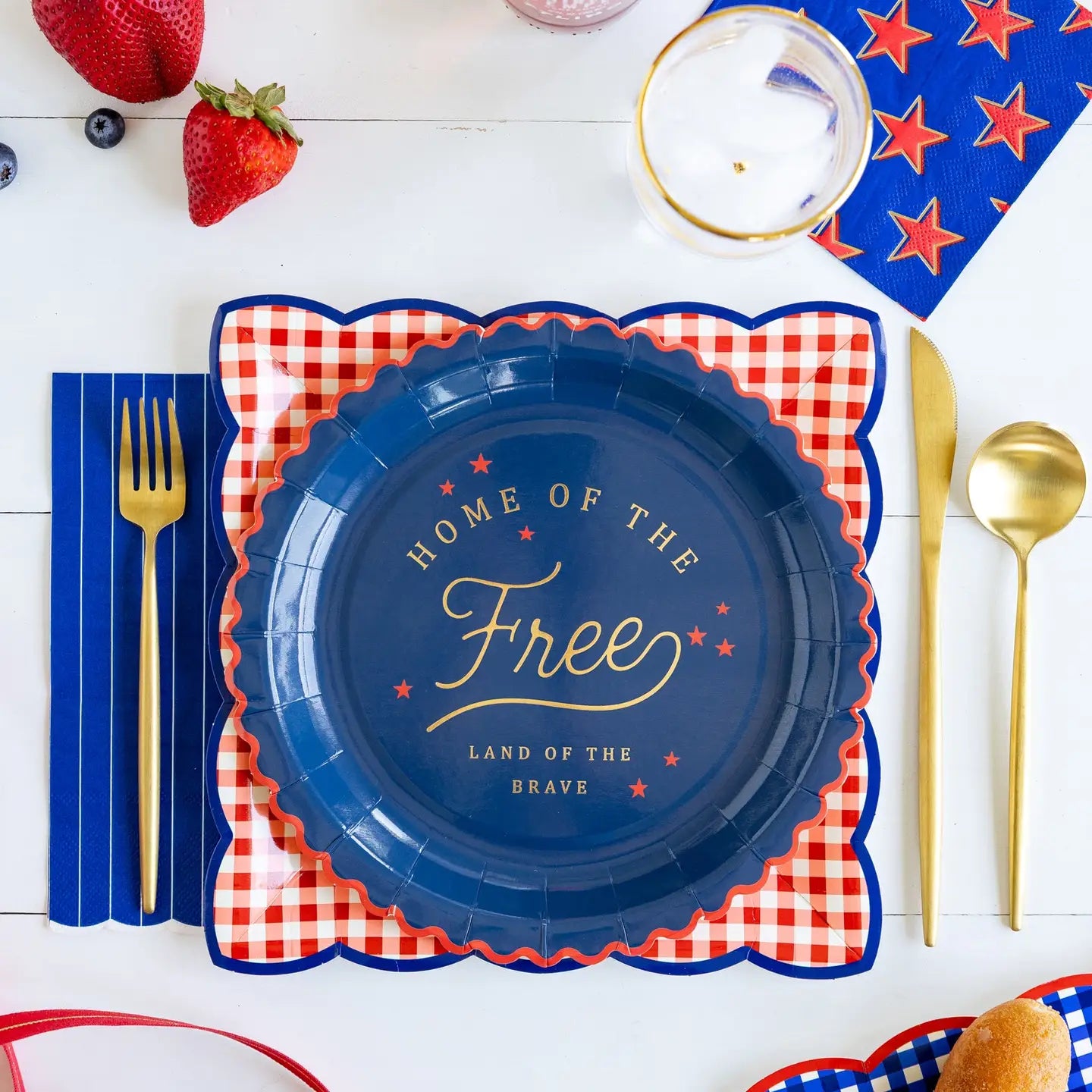 Napkin with 'Home of the Free, Land of the Brave' text on a blue plate with red checkered border, surrounded by gold cutlery and strawberries.